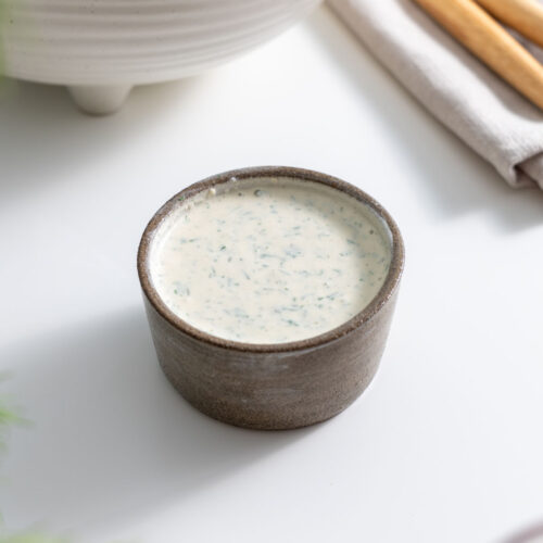 Small bowl of creamy healthy ranch dressing in front of a roasted vegetable bowl on a white surface.