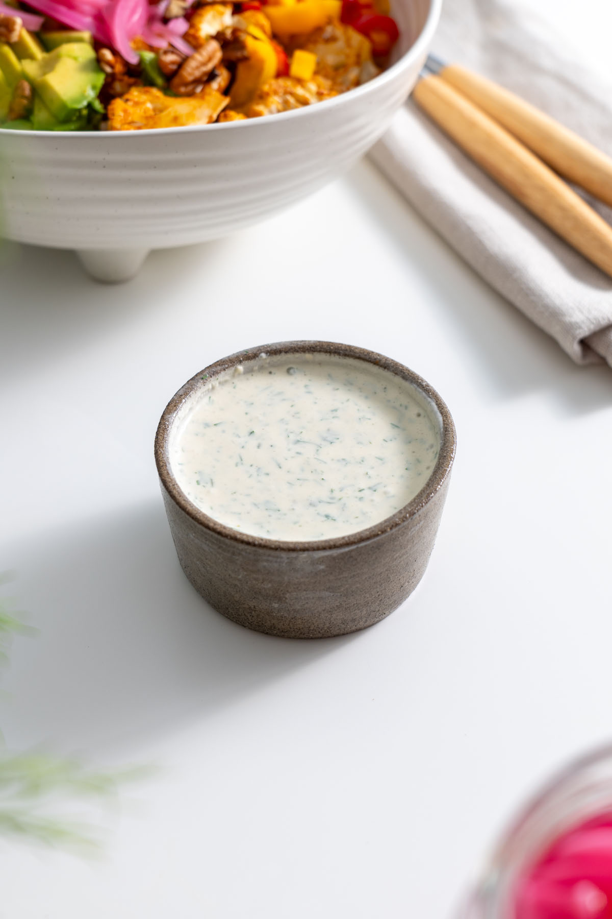 Small bowl of creamy healthy ranch dressing in front of a roasted vegetable bowl on a white surface.