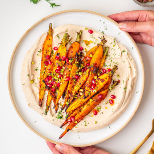 Sarah Cobacho holding a plate of maple roasted carrots with creamy butter bean dip topped with pomegranate seeds, pistachios, and fresh dill