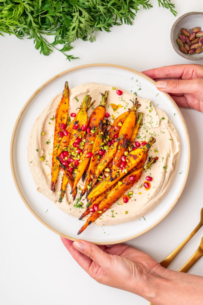 Sarah Cobacho holding a plate of maple roasted carrots with creamy butter bean dip topped with pomegranate seeds, pistachios, and fresh dill