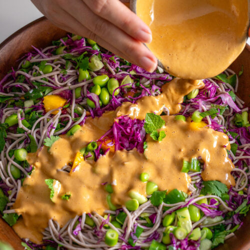 Sarah Cobacho pouring spicy peanut dressing over a colorful soba noodle salad with edamame, cabbage, herbs, and mango.