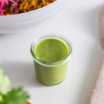 Jar of creamy cilantro dressing on a white surface with a colorful salad bowl in the background.