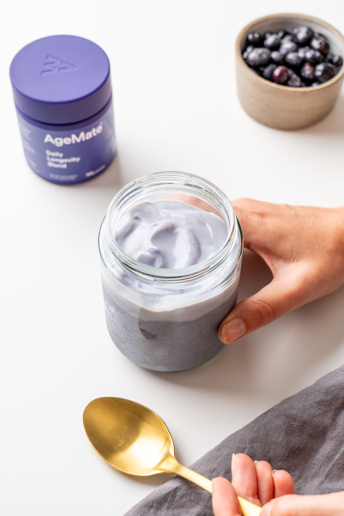 Sarah Cobacho holding a glass jar of creamy blended blueberry oats, with an AgeMate Daily Longevity Blend tub, a bowl of fresh blueberries, and a gold spoon on a white surface