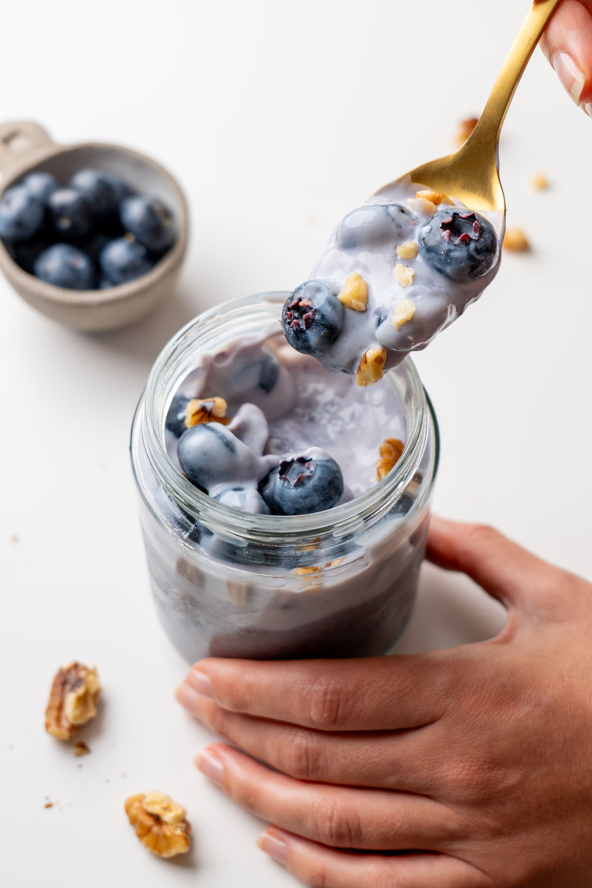 Gold spoon lifting a bite of creamy blended blueberry oats with fresh blueberries and chopped walnuts out of a glass jar, with a small bowl of blueberries in the background