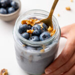 Hand holding a glass jar of blended blueberry oats topped with fresh blueberries and walnuts, with a gold spoon and grey linen napkin on a white surface