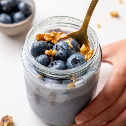 Hand holding a glass jar of blended blueberry oats topped with fresh blueberries and walnuts, with a gold spoon and grey linen napkin on a white surface
