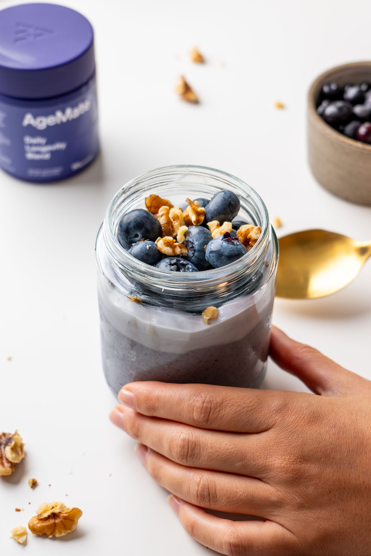 Hand holding a glass jar of blended blueberry oats topped with fresh blueberries and walnuts, with an AgeMate Daily Longevity Blend tub in the background and walnuts scattered on a white surface
