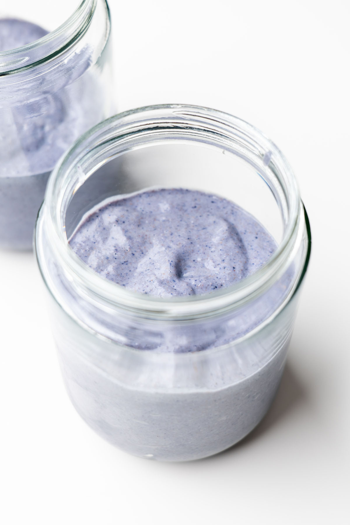 Two glass jars of creamy blended blueberry oats setting on a white surface, ready to chill in the fridge before serving