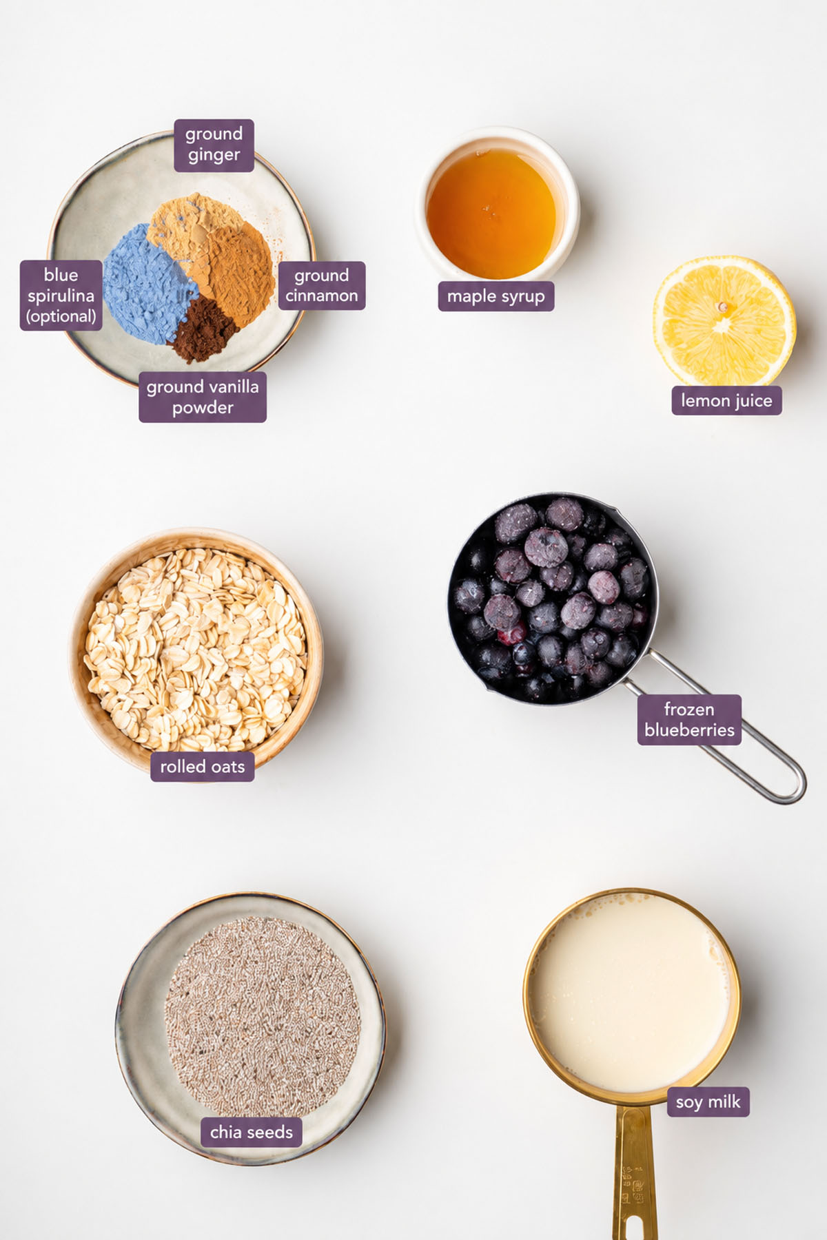 Vibrant ingredients for a blended blueberry oats recipe on a white surface, including rolled oats, chia seeds, frozen blueberries, soy milk, maple syrup, lemon juice, blue spirulina, ground vanilla powder, ground cinnamon, and ground ginger