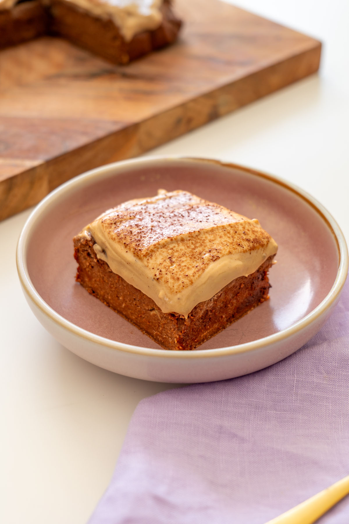 Square slice of brownie baked oats topped with creamy coffee yogurt icing on a pink plate, with a gold fork and another slice on a wooden board in the background.