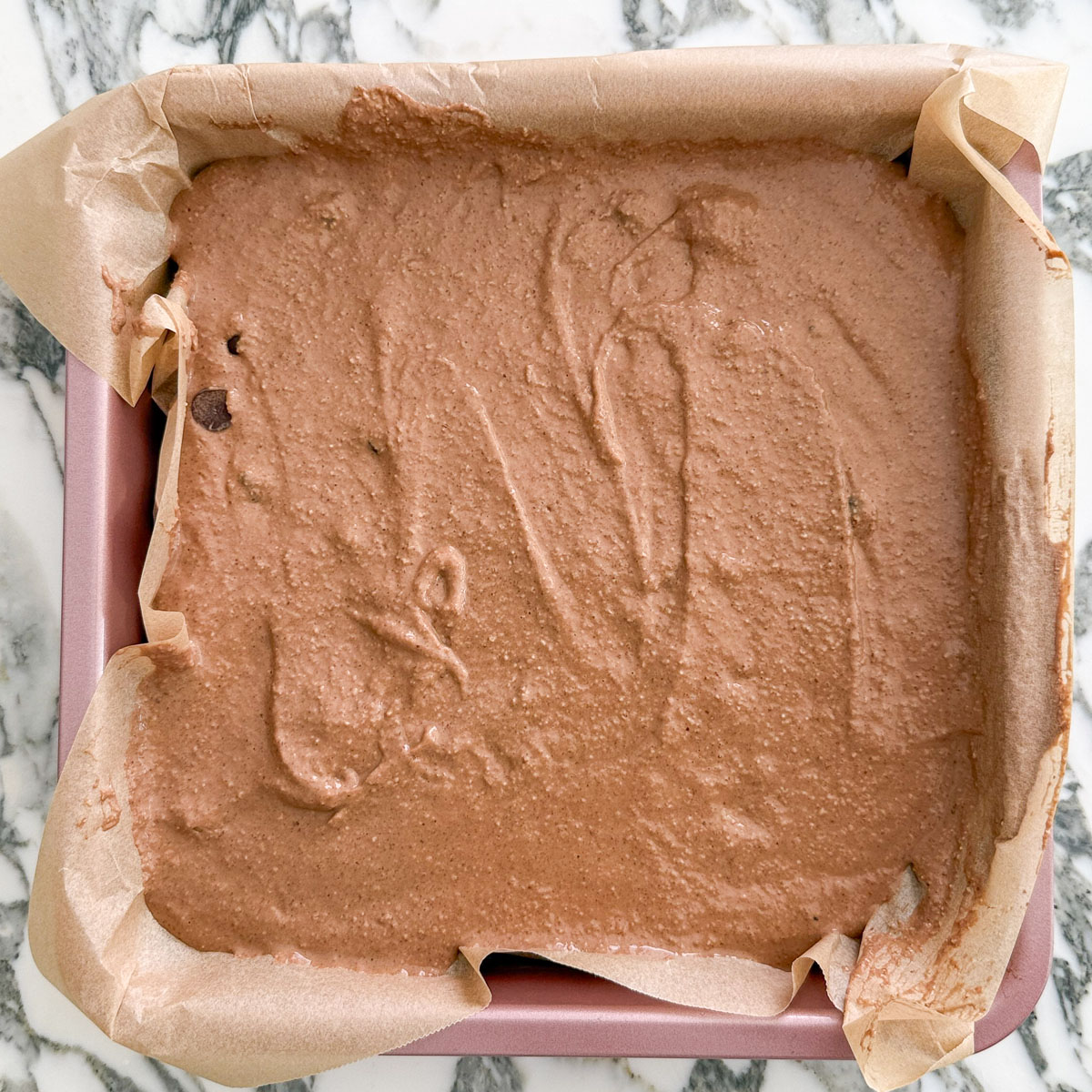 Smooth brownie baked oats batter poured into a parchment-lined square cake pan, ready to bake.
