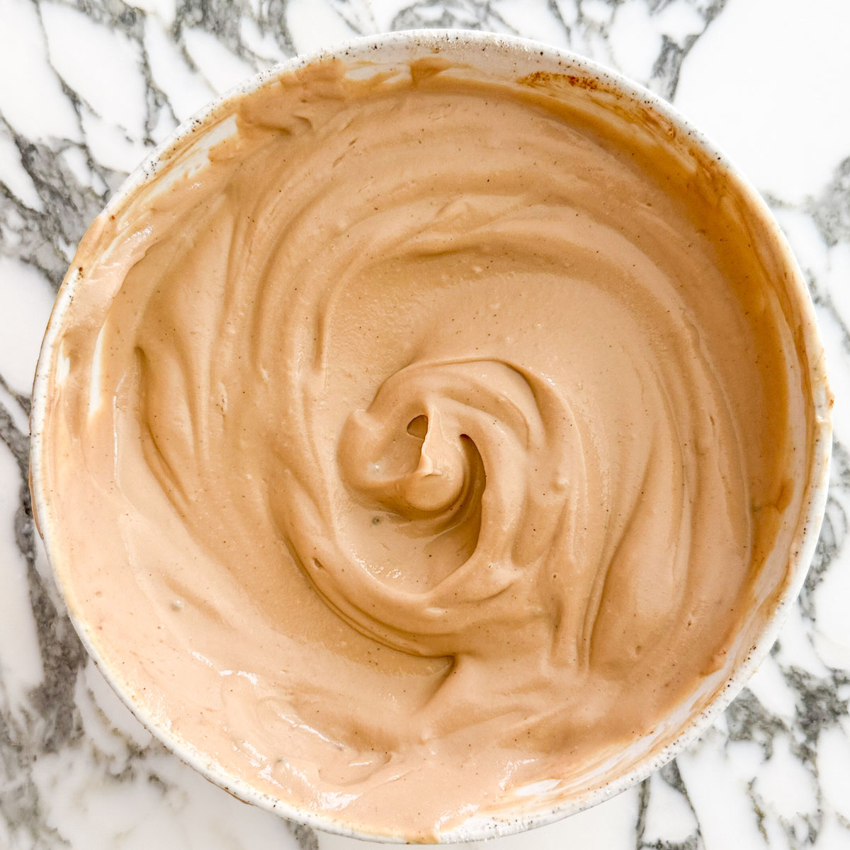 Bowl of smooth coffee yogurt icing made with thick yogurt, vanilla protein powder, and instant coffee on a marble surface.