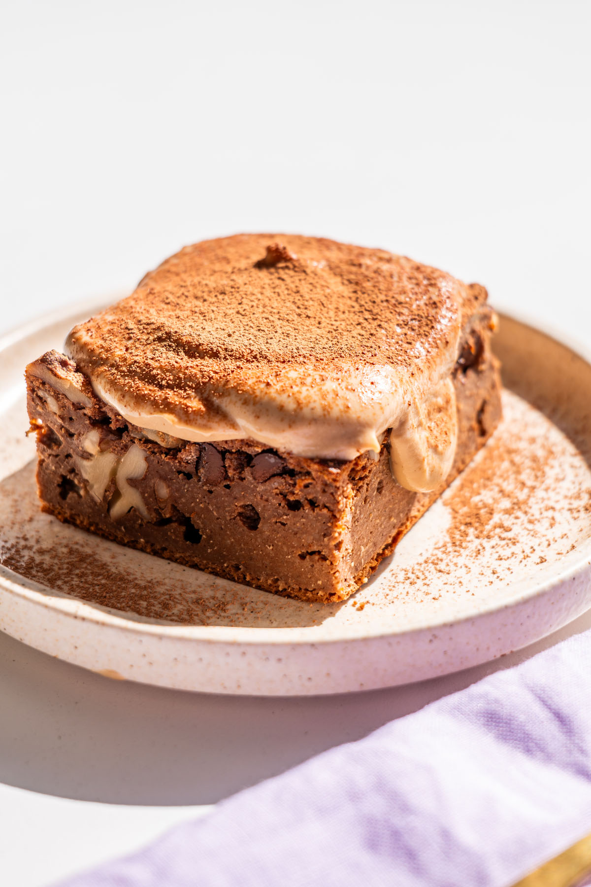 Single slice of brownie baked oats with melting coffee yogurt icing dusted with cacao powder, on a speckled ceramic plate.