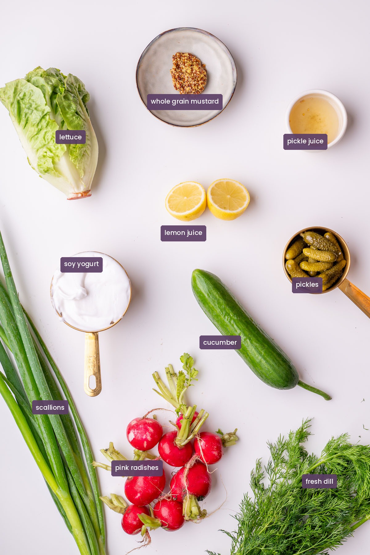 Vibrant ingredients for a crispy shaved potato salad on a white surface, including soy yogurt, lemon, whole grain mustard, pickles, pickle juice, lettuce, cucumber, pink radishes, scallions, and fresh dill