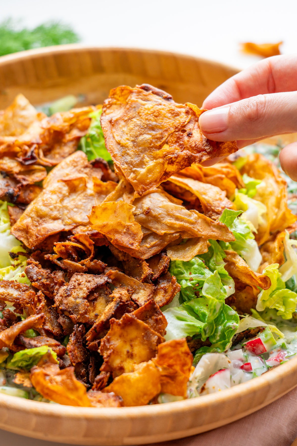 Sarah Cobacho holding a golden crispy shaved potato chip above a wooden bowl of crispy shaved potato salad topped with smoky tofu bacon bits and fresh lettuce