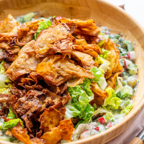 Crispy shaved potato salad in a wooden serving bowl on a marble surface, topped with golden crispy shaved potatoes, smoky tofu bacon bits, fresh dill, and chopped radish in a creamy yogurt-pickle dressing