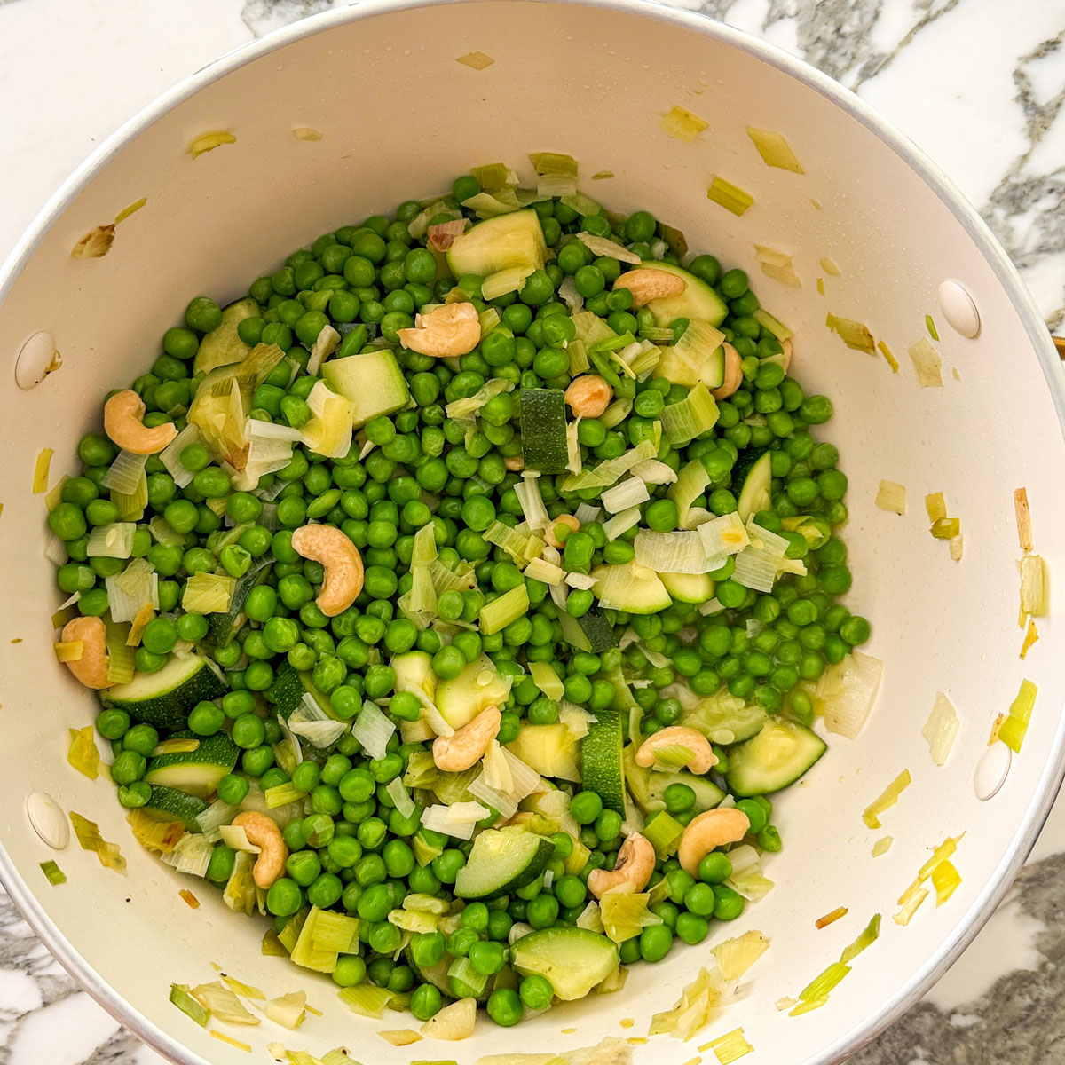 Frozen baby peas, leek, zucchini, and raw cashews in a cream colored pot, partly cooked, ready for the broth