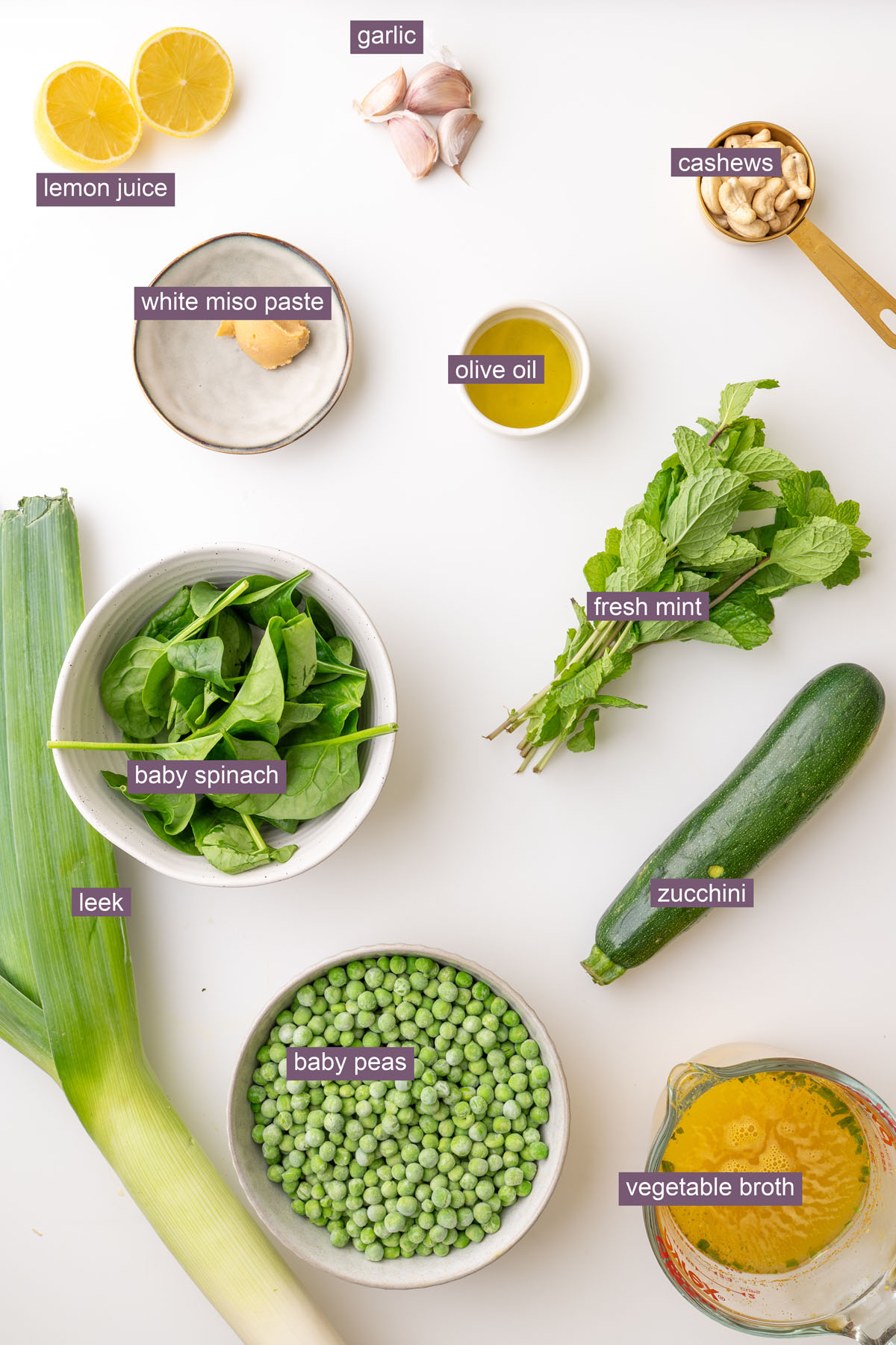 Vibrant ingredients for a sweet pea and mint soup on a white surface, including frozen baby peas, fresh mint, baby spinach, leek, zucchini, garlic, lemon, cashews, white miso, olive oil, and vegetable broth