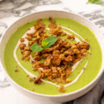 Sweet pea and mint soup in a white speckled bowl, topped with crispy chickpea crumble and a sprig of fresh mint, on a marble surface with a grey linen napkin and a tray of crumble in the background