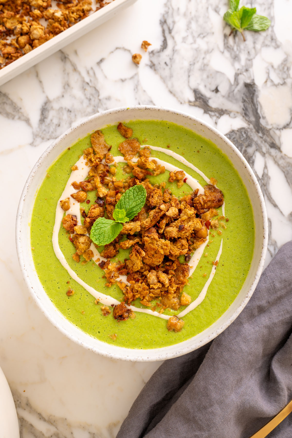 Top-down view of sweet pea and mint soup in a white speckled bowl, topped with chickpea crumble and a mint sprig, with a tray of crumble visible and a golden spoon