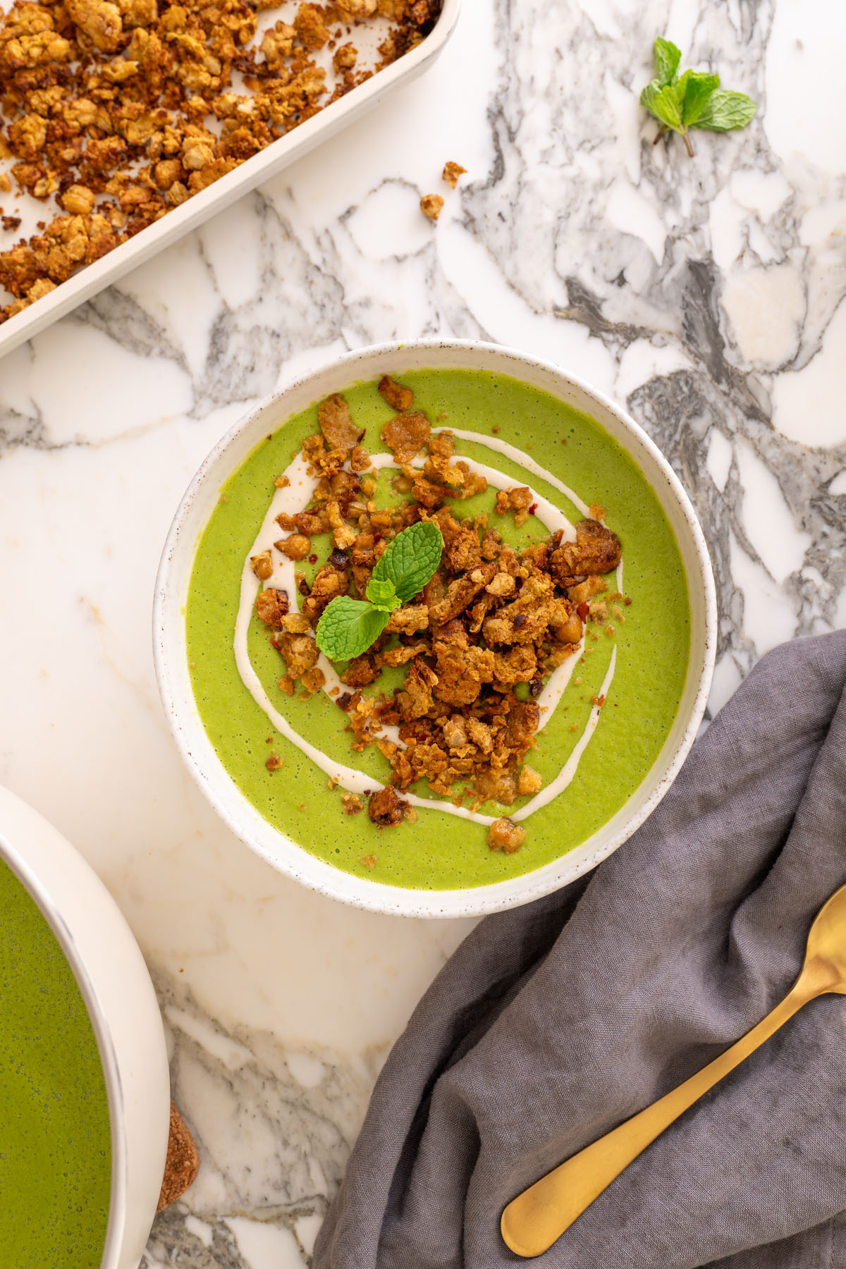 Top-down view of sweet pea and mint soup in a white speckled bowl, topped with chickpea crumble and a mint sprig, with a second bowl partially visible and a golden spoon on the side