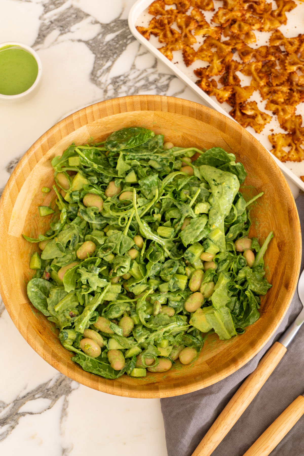 Green salad tossed with green goddess dressing in a wooden bowl, with crispy pasta on a baking tray ready to be added.