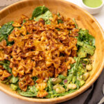 A wooden bowl of viral crispy pasta salad with crunchy pasta, green goddess dressing, chickpeas, avocado, and pistachios on a white surface.