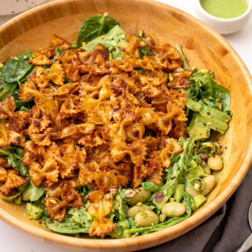 A wooden bowl of viral crispy pasta salad with crunchy pasta, green goddess dressing, chickpeas, avocado, and pistachios on a white surface.