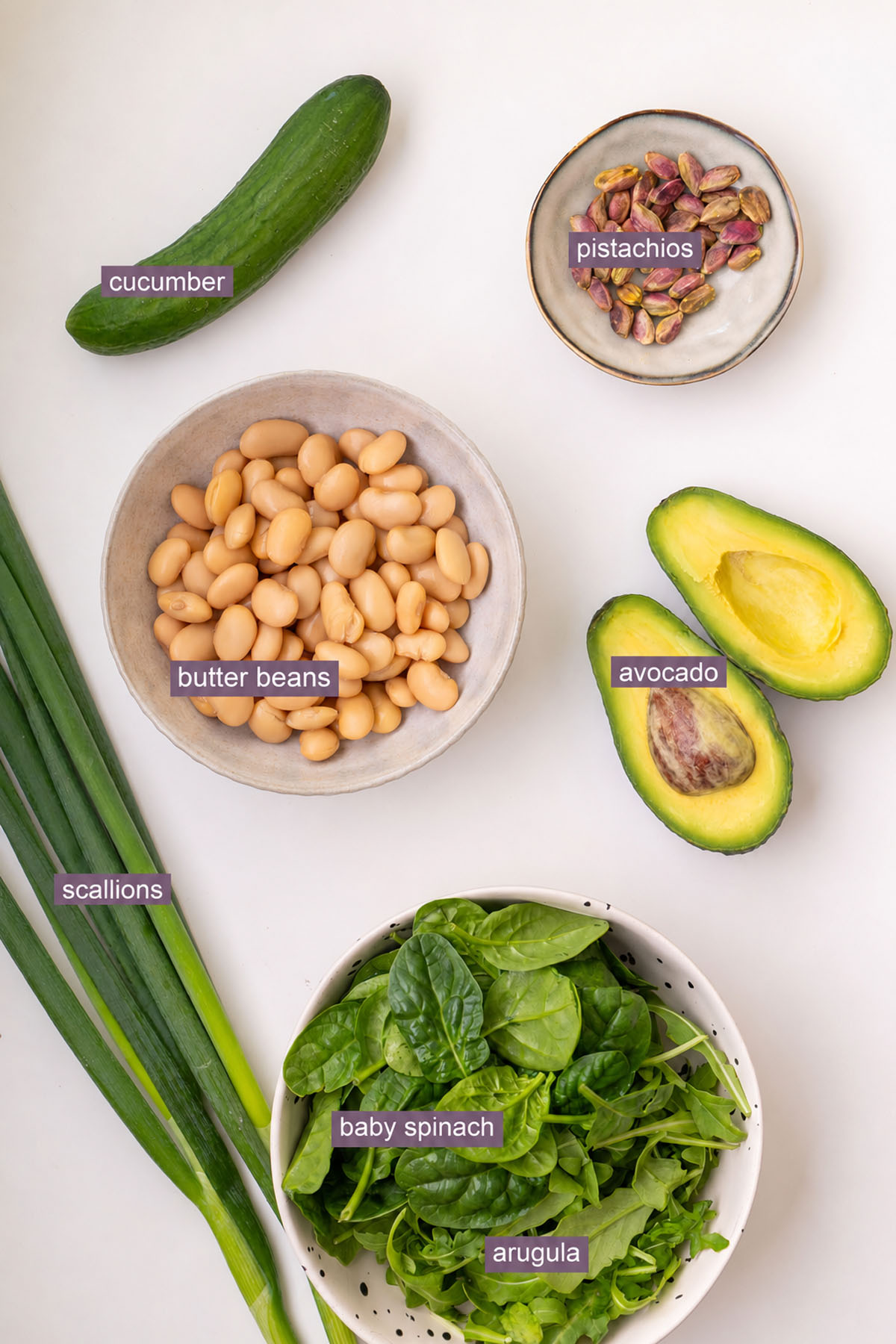 Labeled ingredients for a viral crispy pasta salad, including cucumber, pistachios, chickpeas, avocado, scallions, baby spinach, and arugula on a white surface.
