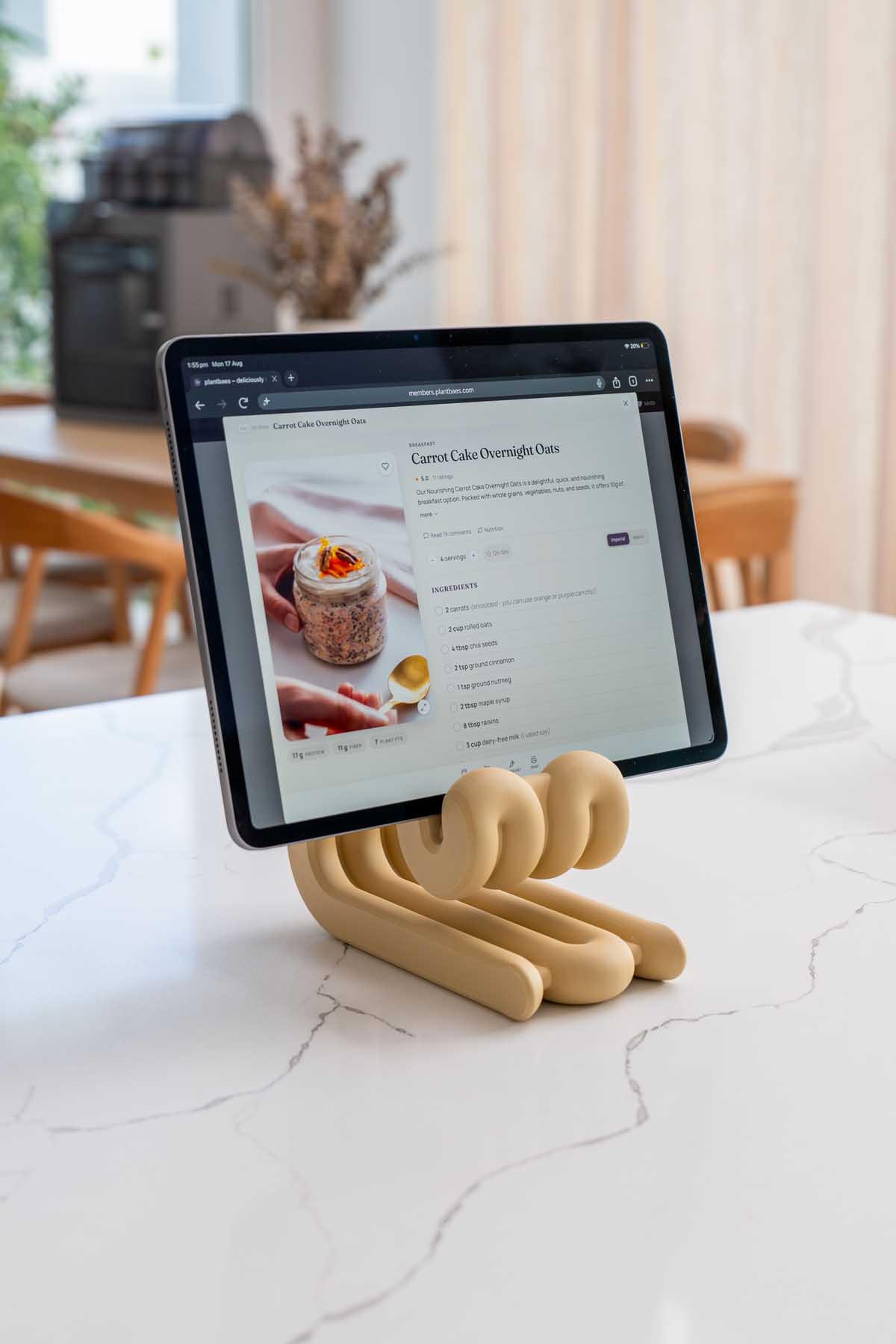 A tablet resting in a 3D printed stand on a marble kitchen bench, open to a plantbaes recipe page