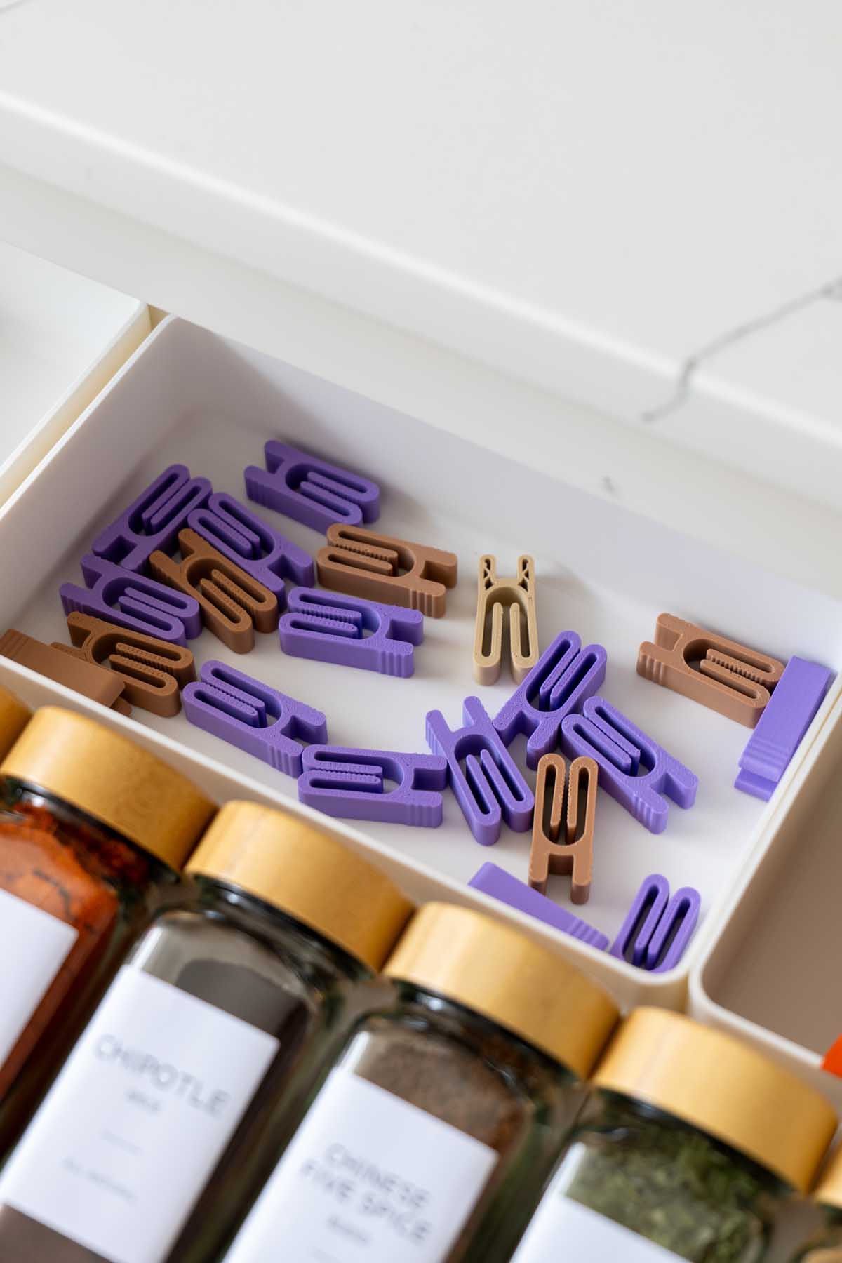 A drawer of 3D printed bag clips in purple and tan sitting beside labeled spice jars