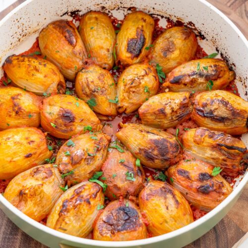 Mediterranean stuffed onions in a pan on a wooden board, baked in tomato sauce and scattered with fresh parsley.