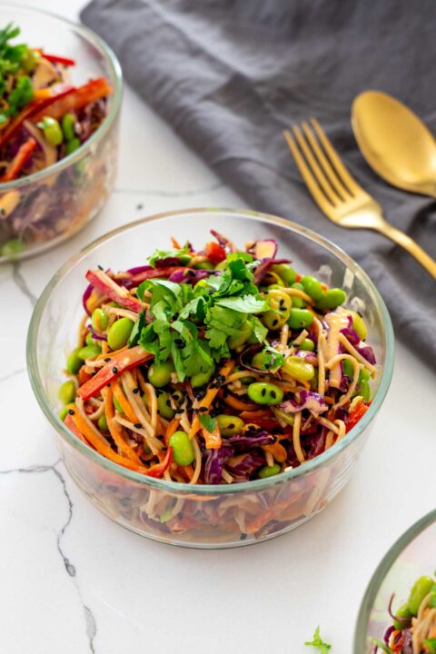 A glass container of spicy peanut noodle salad with soba noodles, edamame, red cabbage, carrot, and bell pepper, topped with fresh cilantro, next to gold cutlery on a grey linen napkin