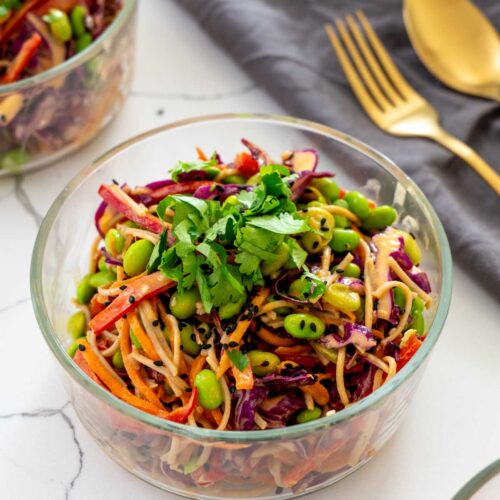 A glass container of spicy peanut noodle salad with soba noodles, edamame, red cabbage, carrot, and bell pepper, topped with fresh cilantro, next to gold cutlery on a grey linen napkin
