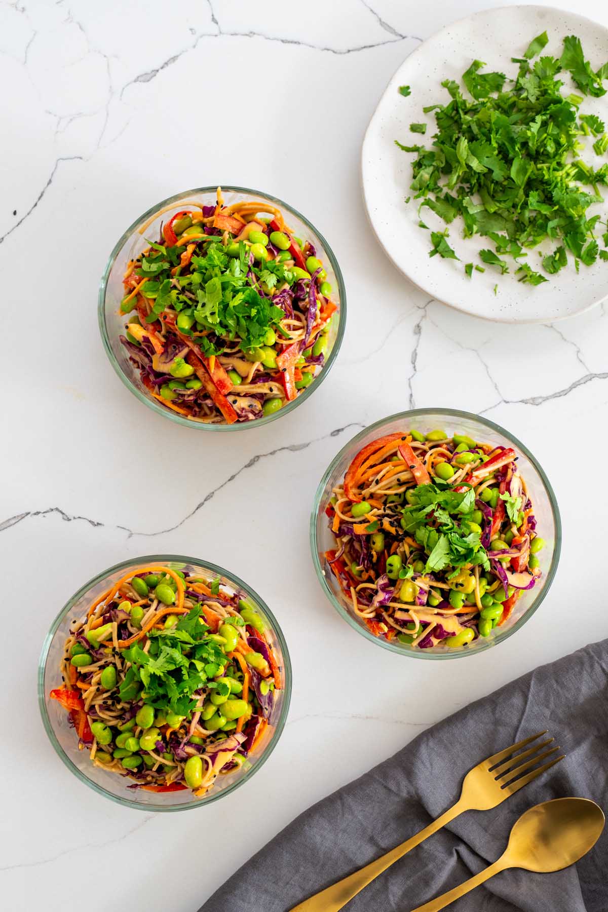 Three glass meal prep containers of spicy peanut noodle salad on a marble surface, each topped with fresh cilantro, beside a plate of chopped cilantro and gold cutlery