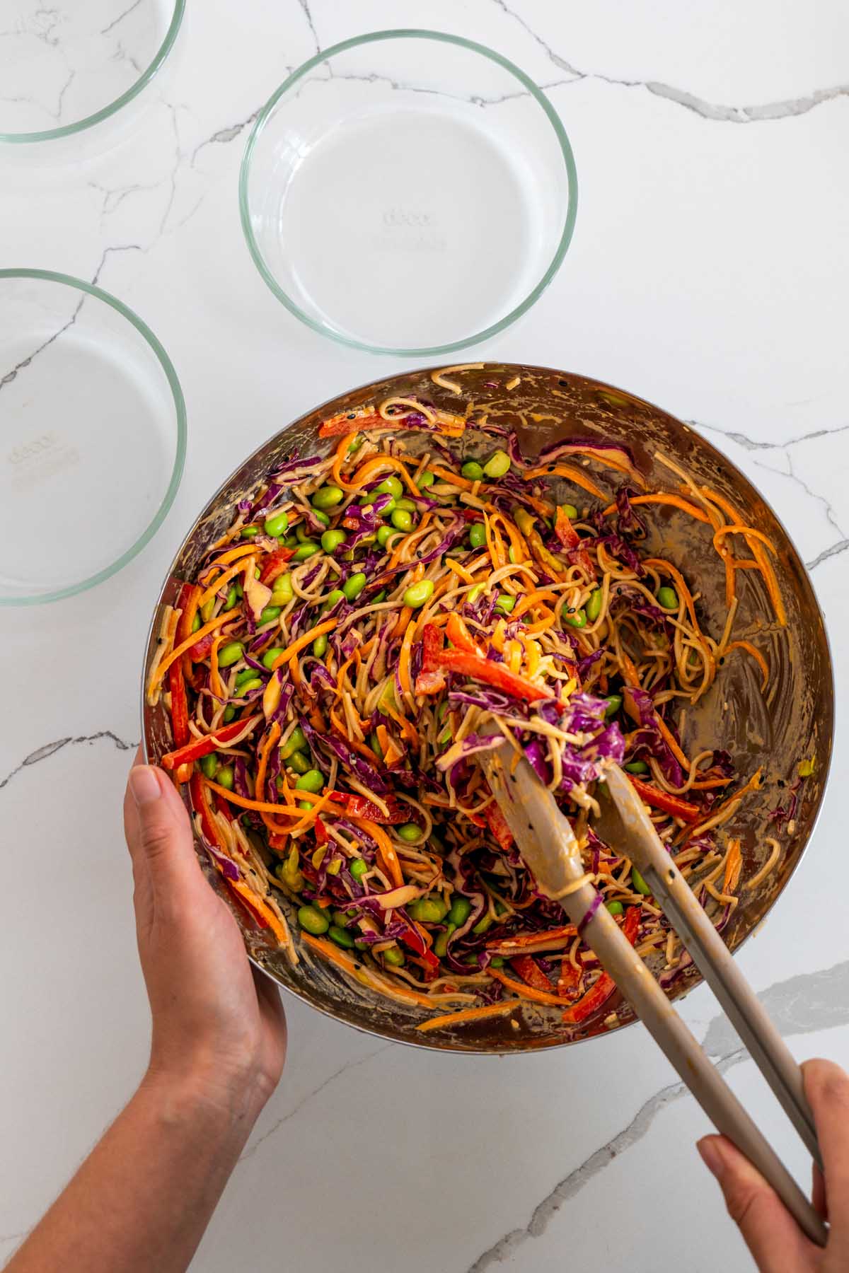 Sarah Cobacho tossing spicy peanut noodle salad in a large glass bowl with wooden tongs, coating the soba noodles, edamame, cabbage, carrot, and bell pepper in the peanut sauce