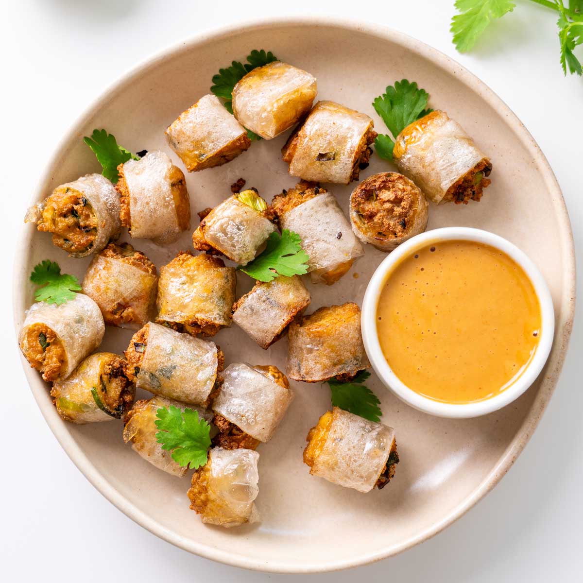 Golden crispy rice paper nuggets piled on a cream plate with a bowl of spicy peanut sauce and scattered fresh cilantro leaves
