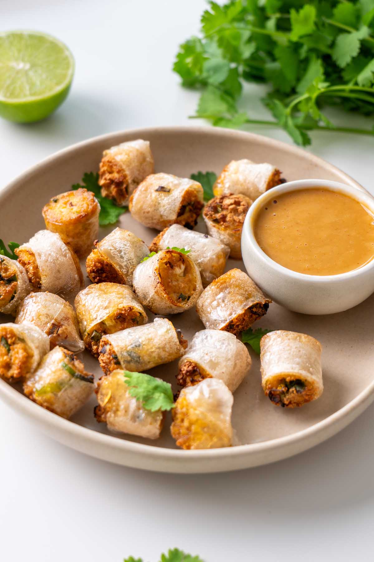 Crispy rice paper nuggets in a shallow cream bowl with a small dish of spicy peanut sauce, fresh cilantro, and a halved lime on a white surface