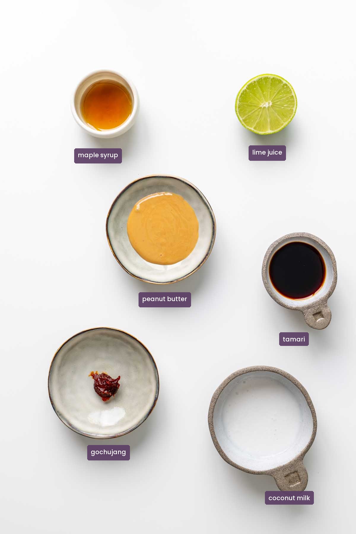 Vibrant ingredients for a spicy peanut dipping sauce on a white surface, including peanut butter, lime juice, tamari, maple syrup, and gochujang