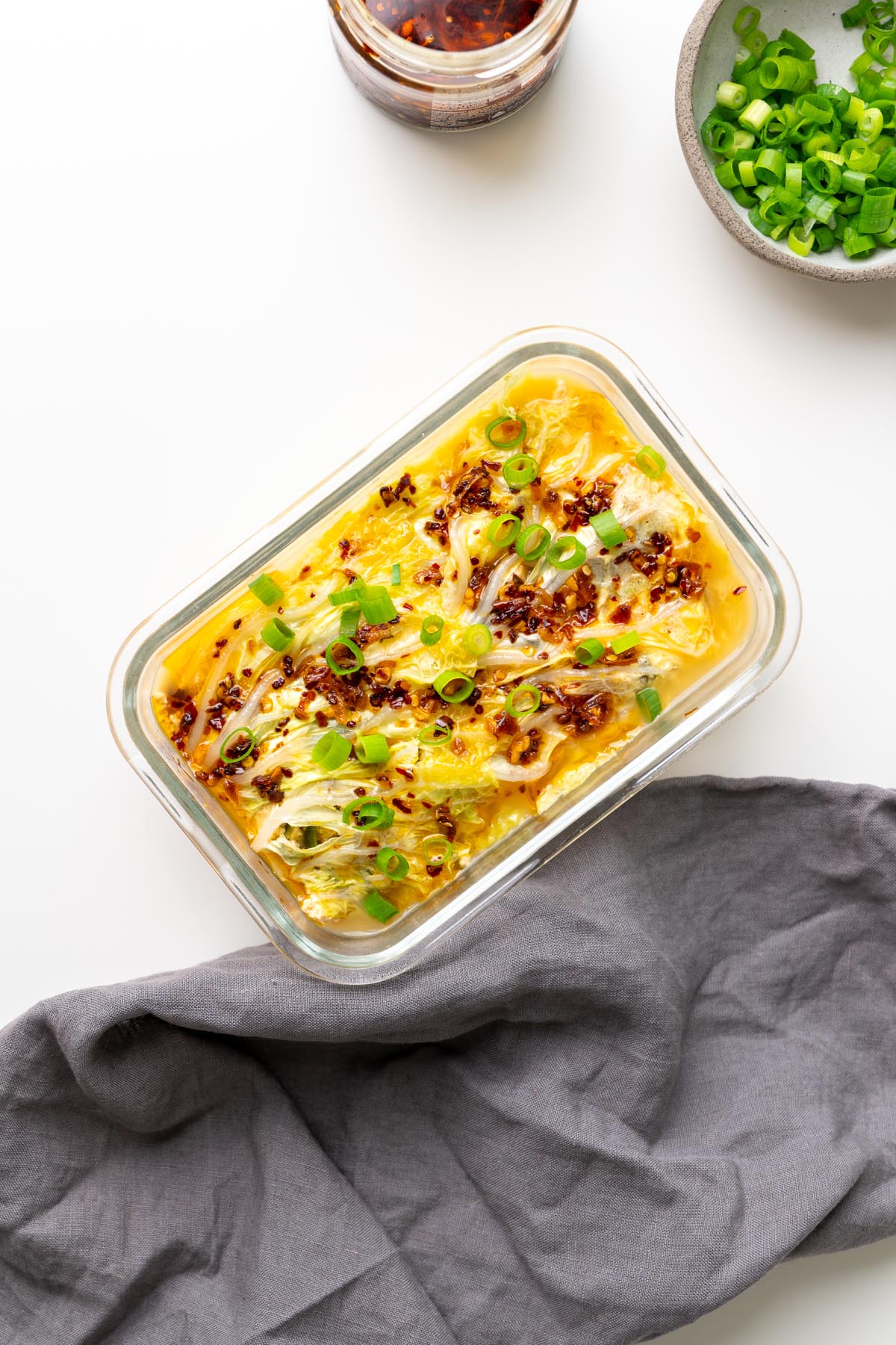 cabbage soup dumpling lasagna in a glass dish on a white surface with a gray linen napkin, topped with crispy chili oil and scallions.