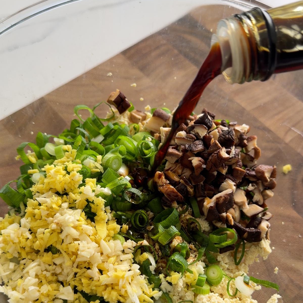 pouring tamari over crumbled tofu, ginger, scallions, and diced shiitake in a glass bowl.