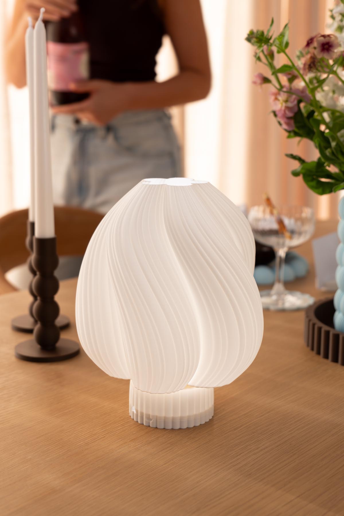 A 3D printed pleated white table lamp glowing softly on an oak dining table, with taper candles and flowers behind it