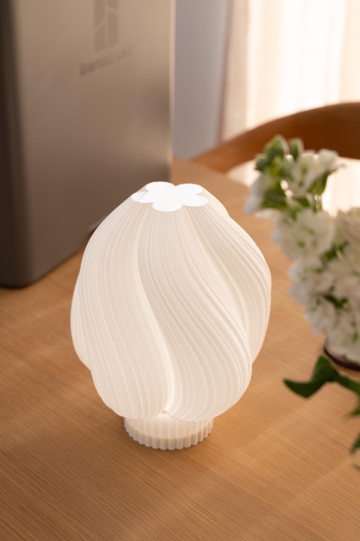 A 3D printed swirled white lamp lit on a dining table next to white flowers, with a Bambu Lab printer box in the background