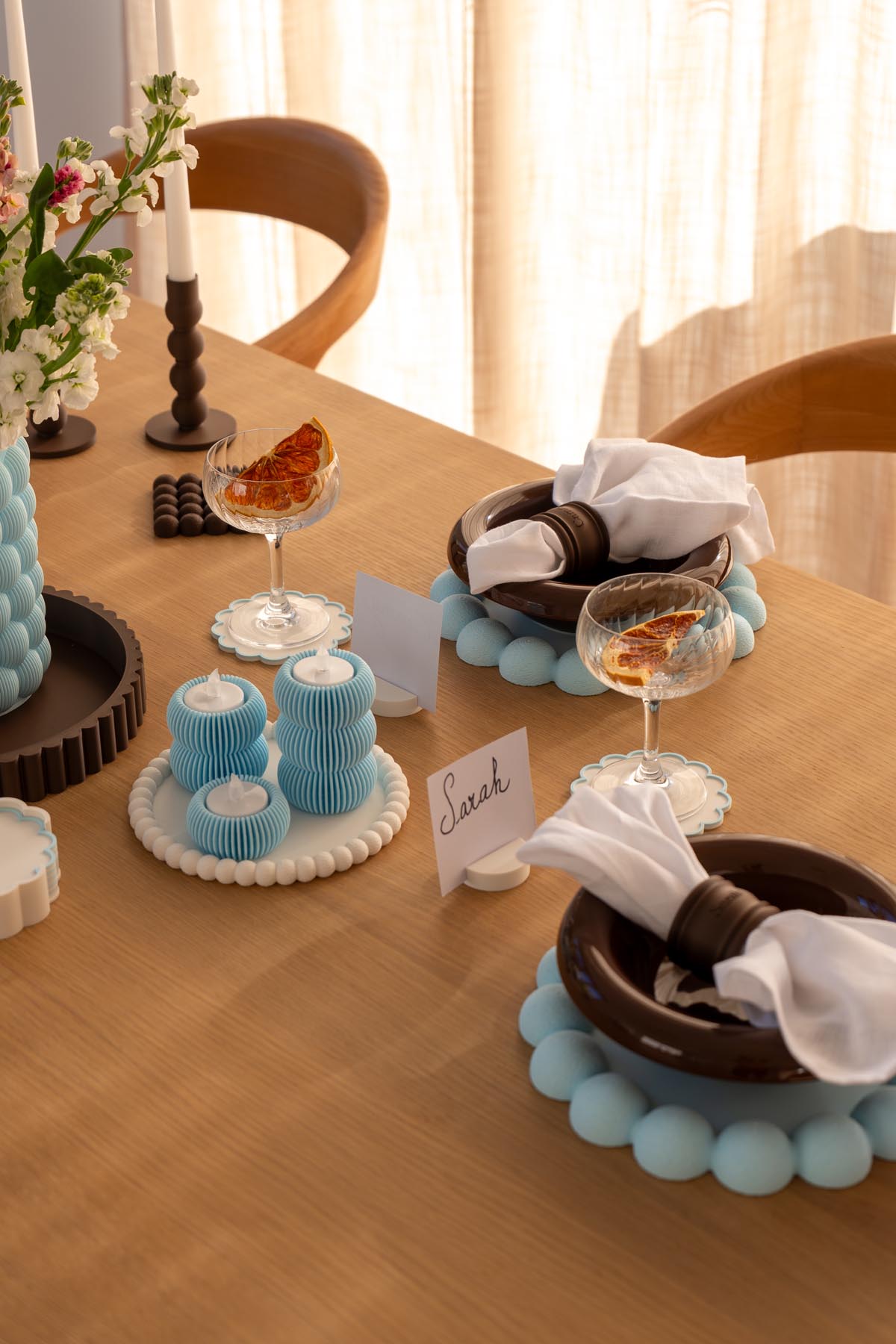 Two place settings with 3D printed blue beaded chargers, brown napkin rings, place card holders and coupe glasses on scalloped coasters