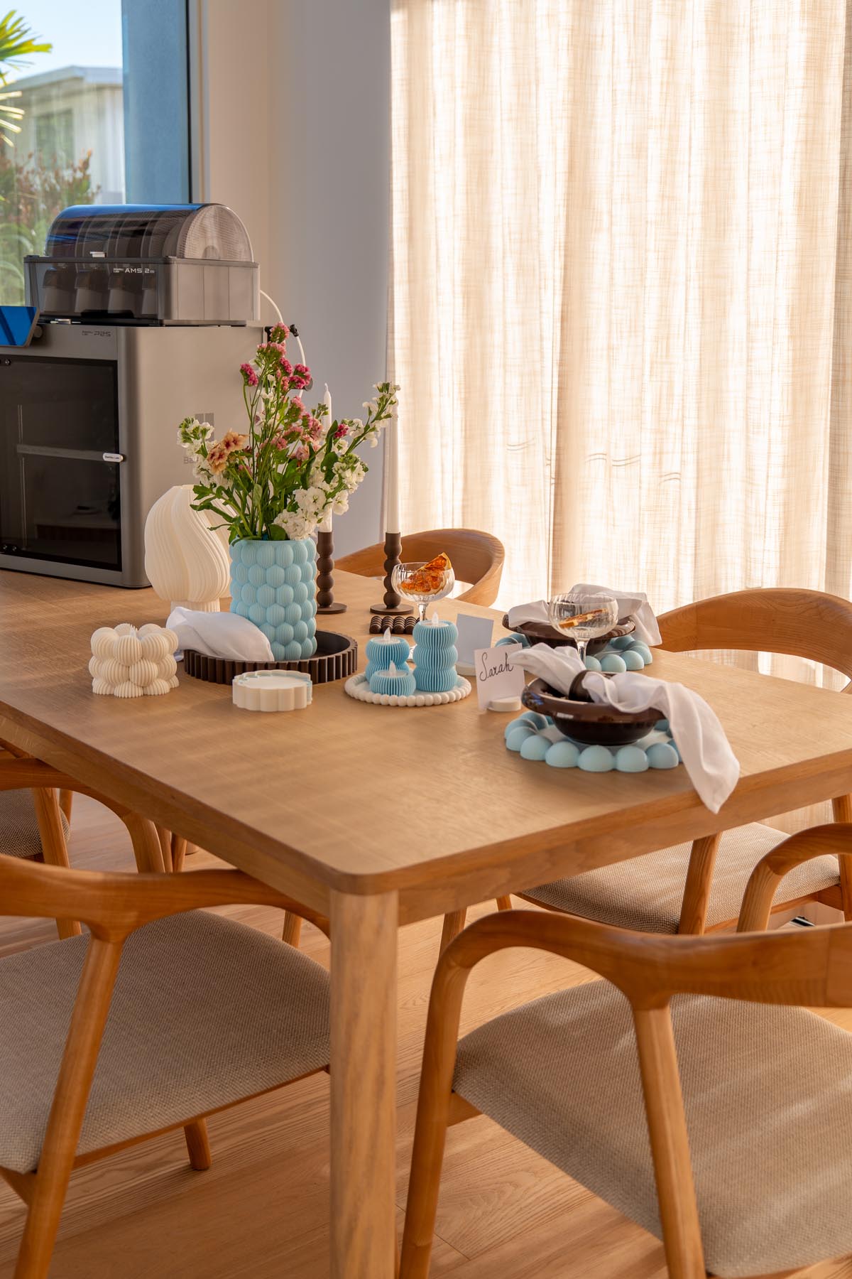 A full dining table styled with 3D printed decor in blue, white and chocolate, with the Bambu Lab printer visible in the corner of the room