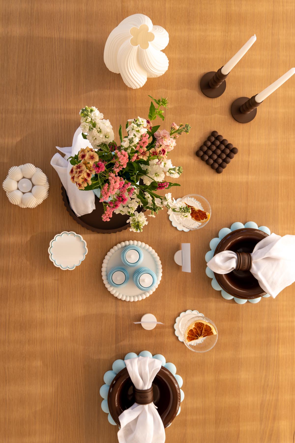 Overhead view of a styled oak dining table with 3D printed chargers, coasters, candle holders, a vase of flowers and a pleated lamp