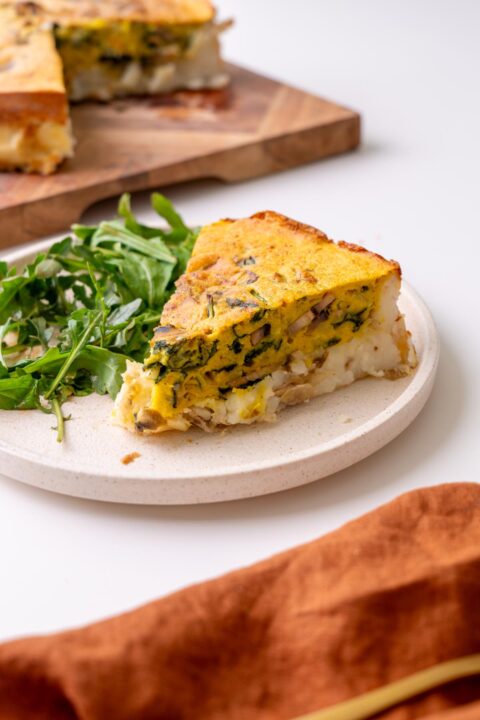 slice of vegan potato crust quiche on a cream plate with arugula, the rest of the quiche on a wooden board behind.