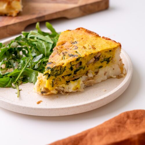 slice of vegan potato crust quiche on a cream plate with arugula, the rest of the quiche on a wooden board behind.